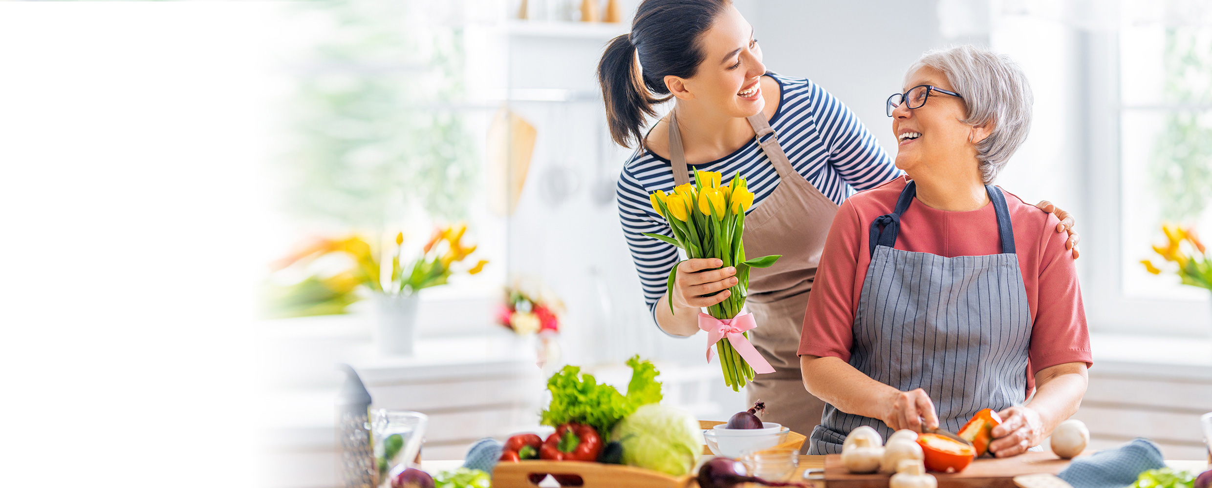 Young woman brings yellow tulips to older woman fighting liver disease, and they smile at each other in white kitchen