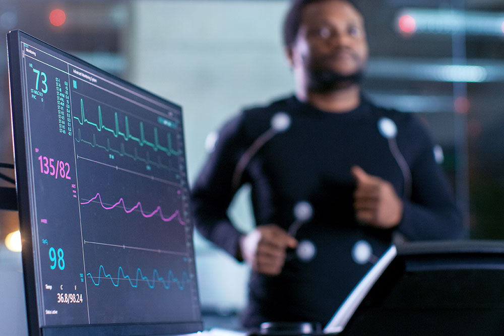 Close-up Shot of a Monitor With EKG Data. Male Athlete Runs on a Treadmill with Electrodes Attached to His Body while Sport Scientist Holds Tablet and Supervises EKG Status in the Background.