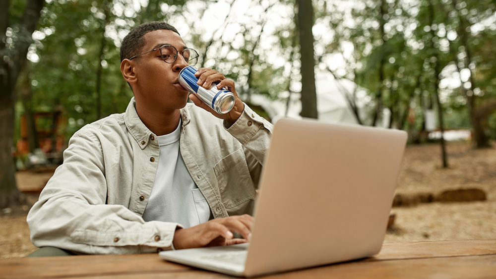 Young man drinks energy drink while sitting at table outside working on computer.